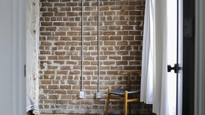 A room with a brick wall, a wooden chair, and a white curtain. - The Grant by Black Swan (Savannah)