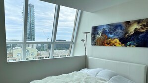 Quarto com uma janela grande, uma cama branca e um quadro na parede.