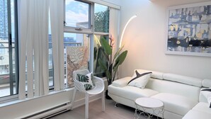 Uma sala de estar moderna com um sofá branco, uma cadeira branca e uma mesa redonda pequena. É visível uma janela grande com cortinas, e uma planta está colocada perto da janela.