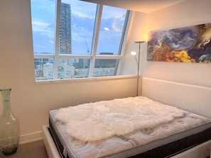 Enjoy a city view from the large window, featuring a tall building and a crane. The room offers a comfortable bed and a floor lamp, accompanied by a unique abstract painting.