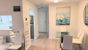Uma sala de estar moderna com uma mesa de jantar de vidro, cadeiras brancas, um frigorífico embutido e um quadro com barcos a vela na parede.