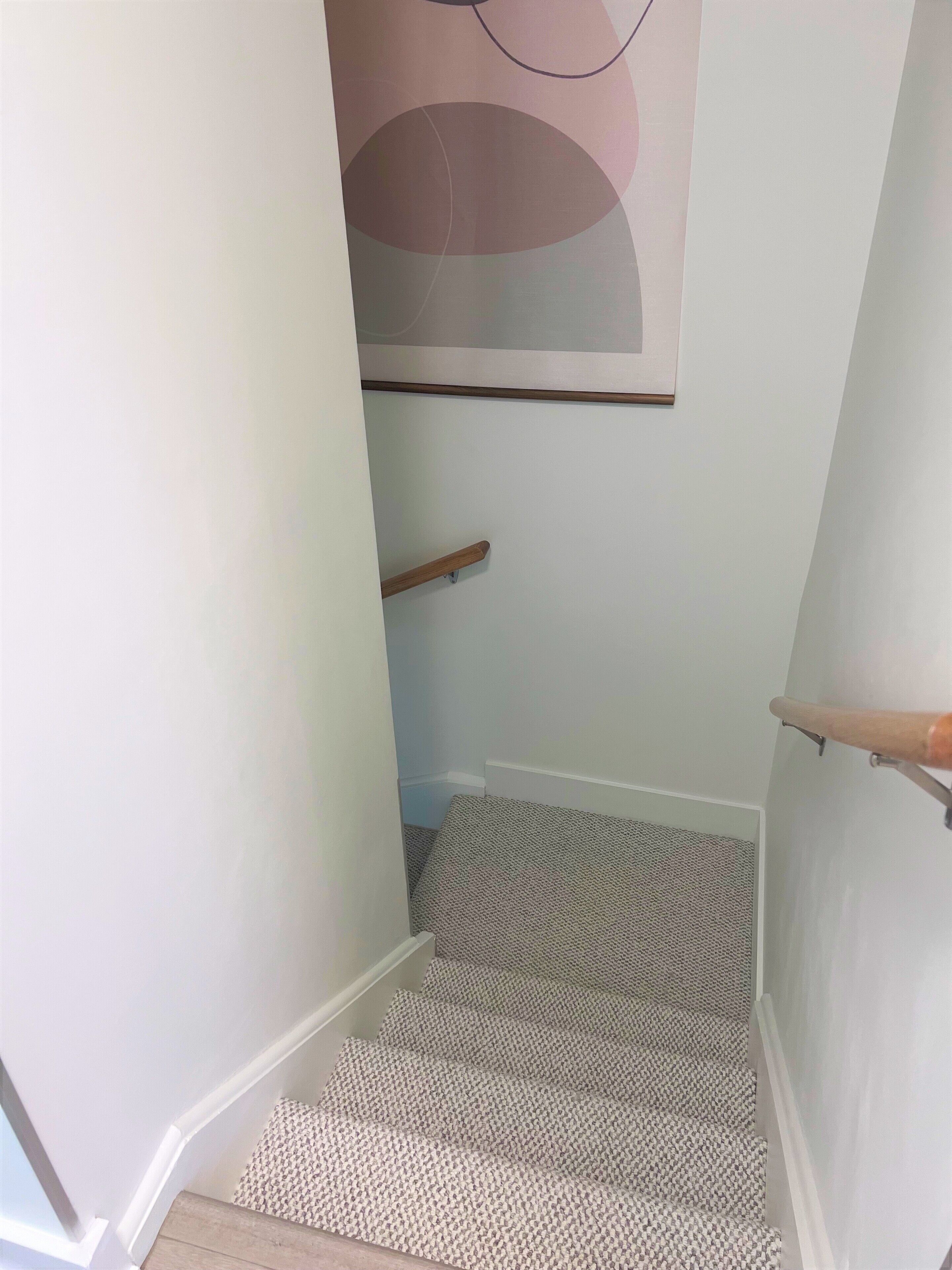 A staircase with patterned carpet and wooden handrails, leading to a room with a plain wall and a large abstract painting.