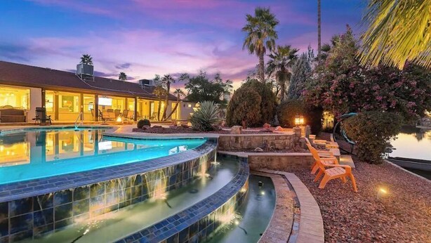 A swimming pool with a curved edge, surrounded by a landscaped garden with palm trees and flowering plants. - Lakefront Resort Villa - Heated Infinity Pool & 8-Seat Spa - Kayaks - 4BR (Mesa)