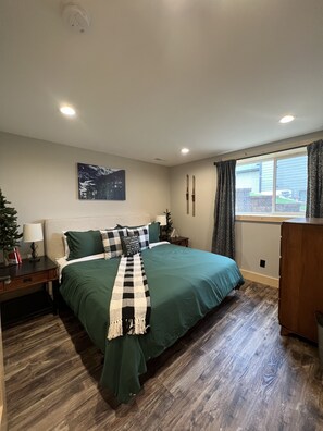 Une chambre avec un grand lit, une table de chevet, une commode, une fenêtre avec des rideaux, un tableau accroché au mur et un luminaire au plafond.
