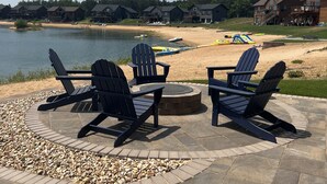 Un espace extérieur aménagé avec des chaises Adirondack et un foyer, offrant une vue sur un lac et une plage de sable, avec des chaises de plage et des parasols.