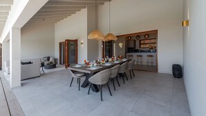 A spacious dining area with a long table set for six, surrounded by wicker chairs. A kitchen with bar stools is visible in the background. - Brand new luxurious villa with bay and mountain view (Curaçao)