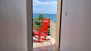 A hallway leading to a balcony with a red rocking chair, a view of the ocean, and a white railing.