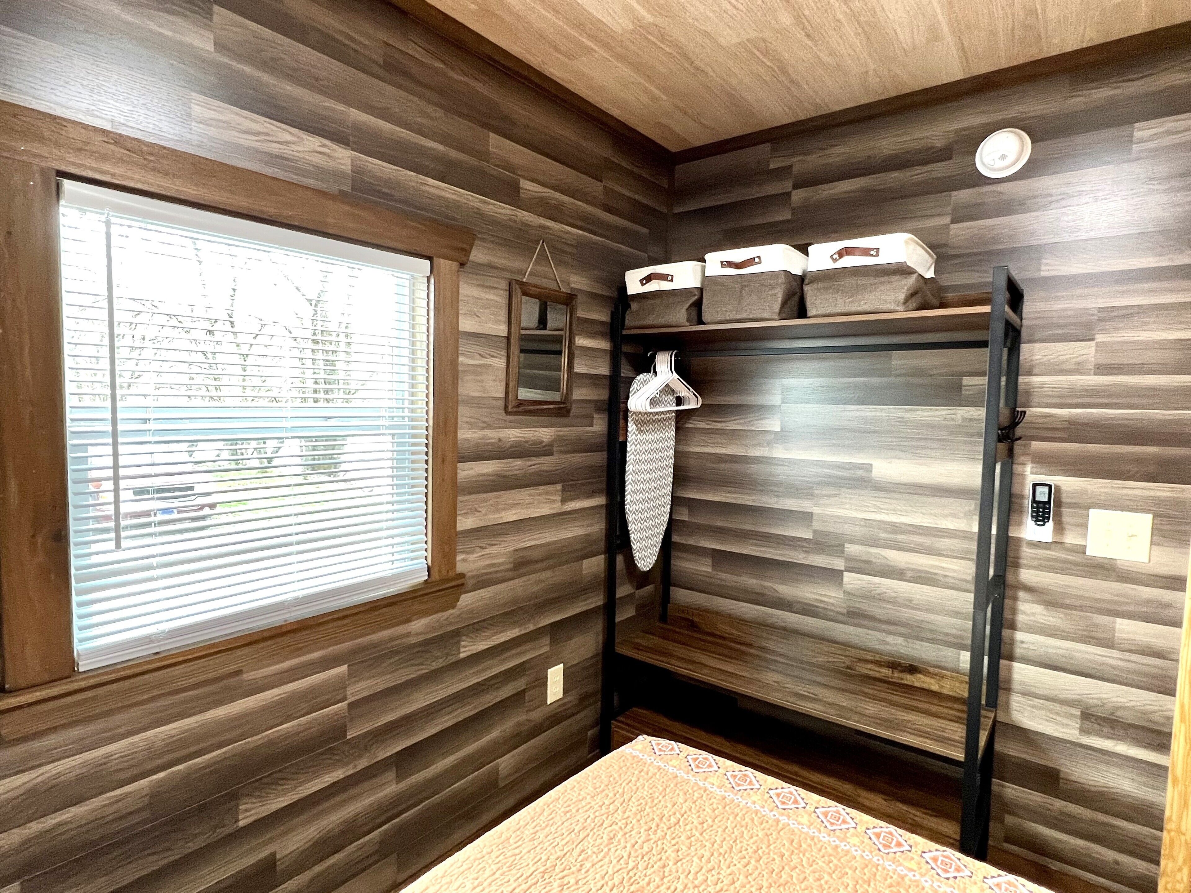 A room with wooden panel walls, a window with blinds, a hanging garment, a bed, and a bedside table.