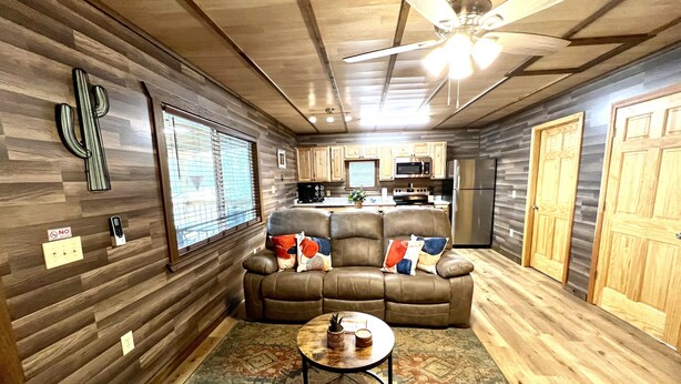 Un espace de vie compact comprenant un canapé, une petite table, un ventilateur de plafond et un coin cuisine avec des meubles et des appareils électroménagers en bois.
