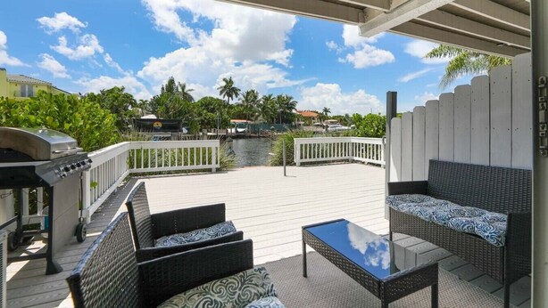 Une terrasse extérieure couverte, aménagée avec des meubles en osier, un barbecue et offrant une vue sur une étendue d’eau et des bâtiments en arrière-plan.