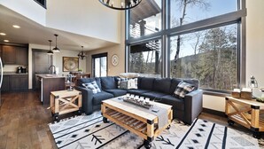 A spacious living room with a large window, a dark-colored sofa, a wooden coffee table, and a kitchen area in the background. - Restful Retreat with Panoramic Views, Hot Tub, and Centrally Located to Deadwood (Lead)