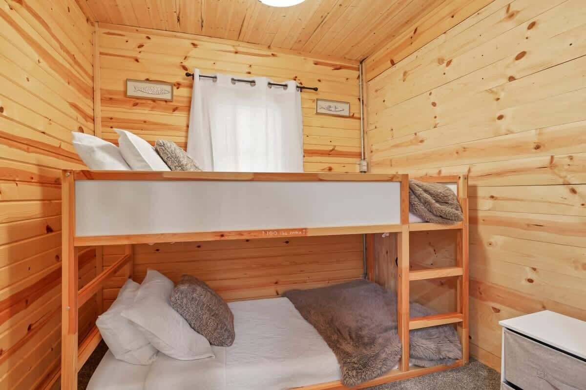 A wooden cabin room with bunk beds, a small desk, and a window with curtains.