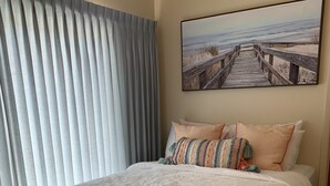 Un dormitorio con una cama grande, un banco, un cuadro de una playa y una ventana con persianas.