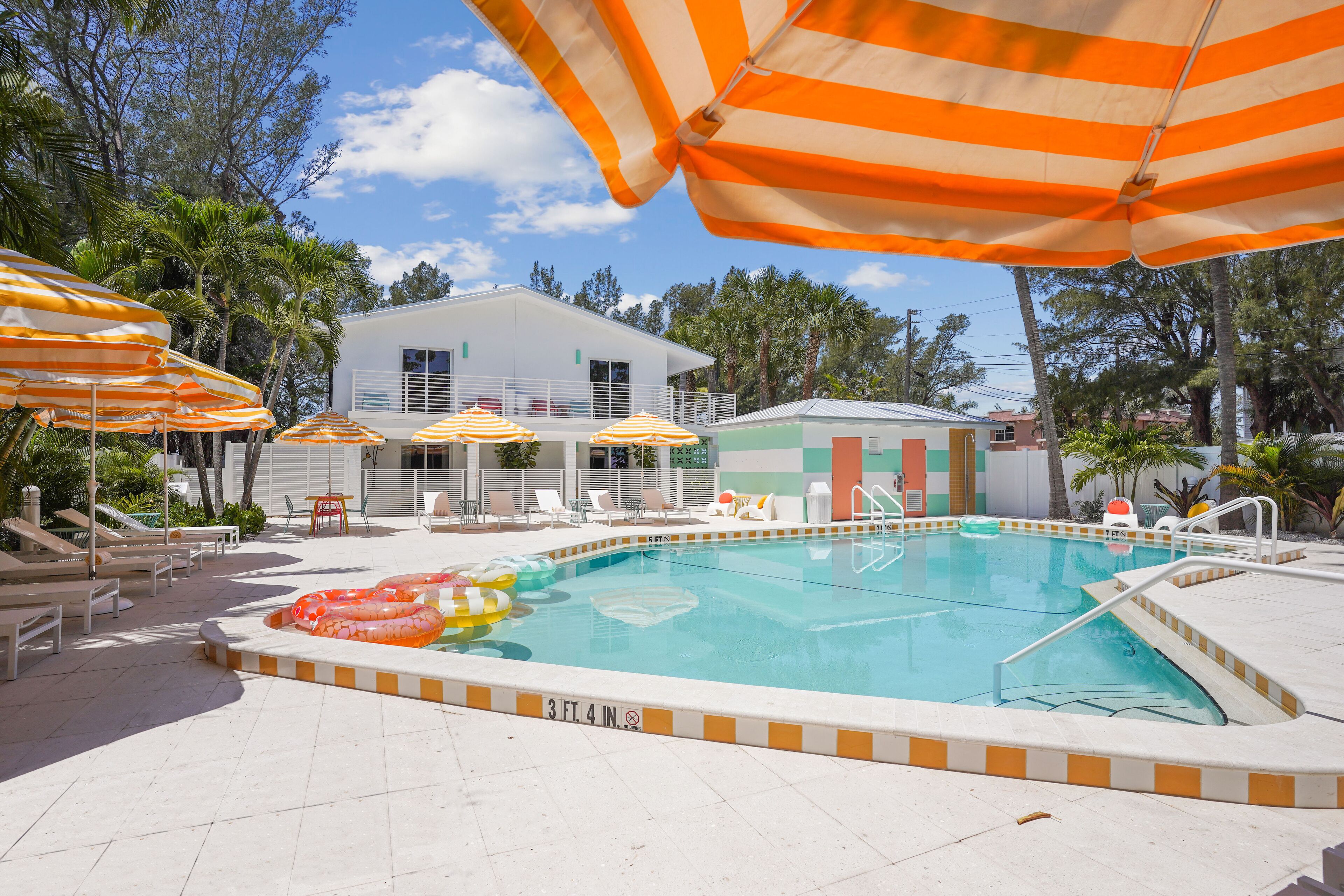 A swimming pool with lounge chairs and striped umbrellas, surrounded by palm trees and a building in the background.