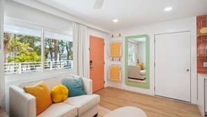 Una sala moderna con un sofá blanco, un ventilador de techo, un ventanal con vistas a palmeras y una puerta con espejo y perchas para toallas.