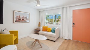 Una sala de estar moderna con un sofá blanco, un sillón amarillo y una mesa de centro blanca. Se observa un cuadro enmarcado en la pared, un ventilador de techo y una ventana con cortinas.