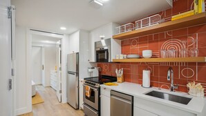 A modern kitchen with white cabinetry, a stainless steel refrigerator, a built-in oven, and a dishwasher. There are orange and white patterned tiles on the backsplash, and a wooden shelf above the sink. - Mello on the Beach - Shore Enuff (Holmes Beach)