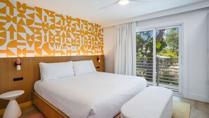 Una habitación de hotel moderna con una cama grande, una mesita de noche pequeña, un ventilador de techo y vistas al exterior a través de un ventanal.