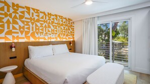 A modern hotel room with a large bed, a bedside table, a small stool, a ceiling fan, and a window with a view of greenery. - Mello on the Beach - Gone Coastal (Holmes Beach)