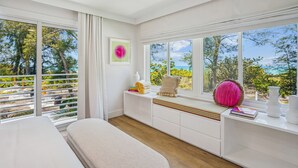 A bright, modern bedroom with large windows, a white bed, and a view of trees and a beach. - Mello on the Beach - Slow M'Ocean (Holmes Beach)