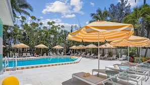 A poolside area with striped umbrellas, lounge chairs, and a swimming pool. - Mello on the Beach - Slow M'Ocean (Holmes Beach)