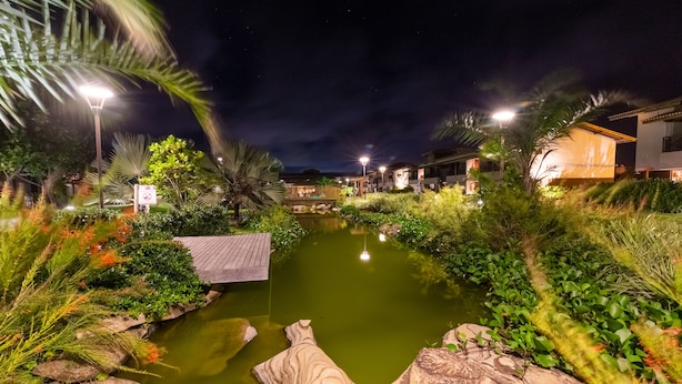 A nighttime view of a landscaped area with a pond, surrounded by greenery and illuminated by street lamps. - Areia Azul Aparts Brazil - Imbassai Ykutiba (Mata de São João)