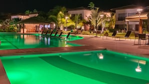 A nighttime pool area with green lighting, lounge chairs, and a building with balconies. - Areia Azul Aparts Brazil - Imbassai Ykutiba (Mata de São João)