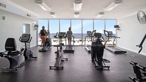 Ett gym med löpband, motionscyklar och en roddmaskin, med utsikt över en strand.
