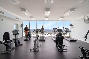 A gym with treadmills, stationary bikes, and a rowing machine, overlooking a beach.