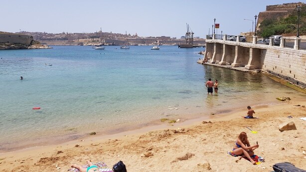 Ein Strand mit klarem Wasser, einem Steinsteg und Booten in der Ferne.