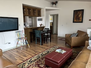 Sala de estar com sofá de couro marrom, um banquinho vermelho e uma TV fixada na parede. Há uma área de jantar com mesa e cadeiras de madeira, e uma cozinha com armários de madeira e uma geladeira.