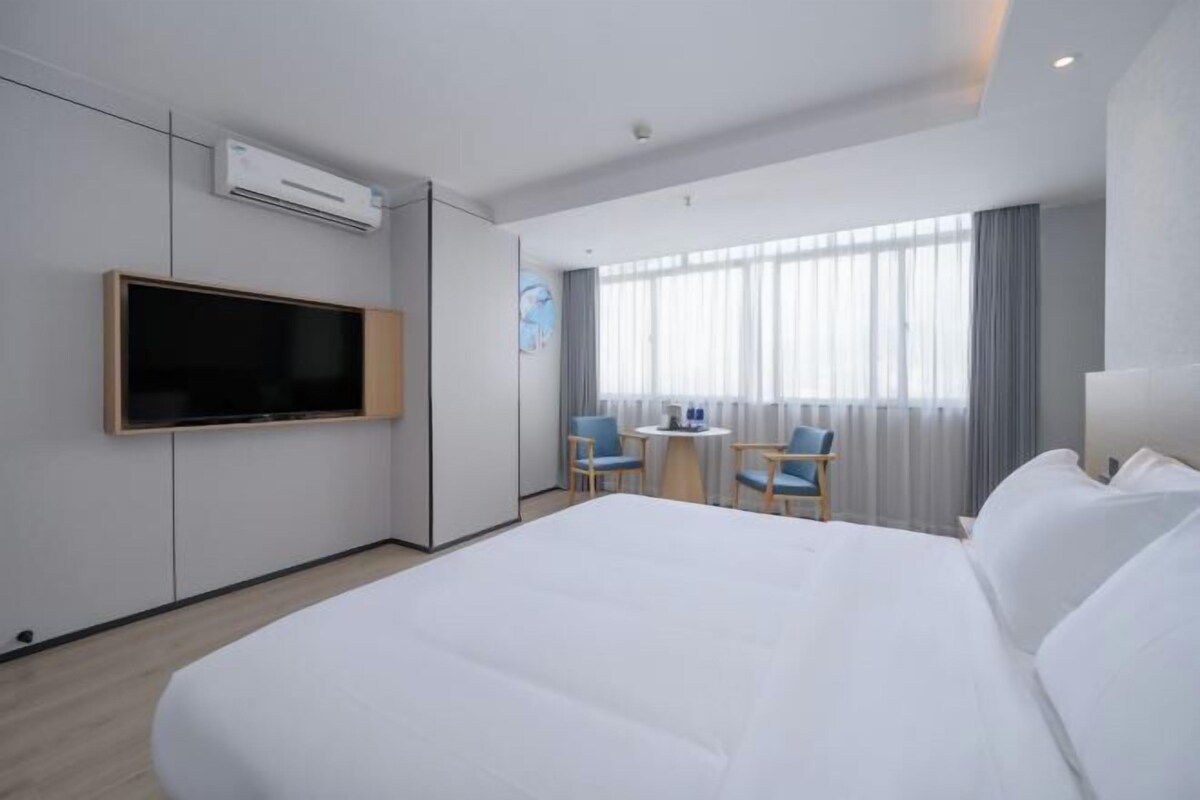 A hotel room with a large bed, a flat-screen TV mounted on the wall, a small table with two chairs, and a large window with curtains.