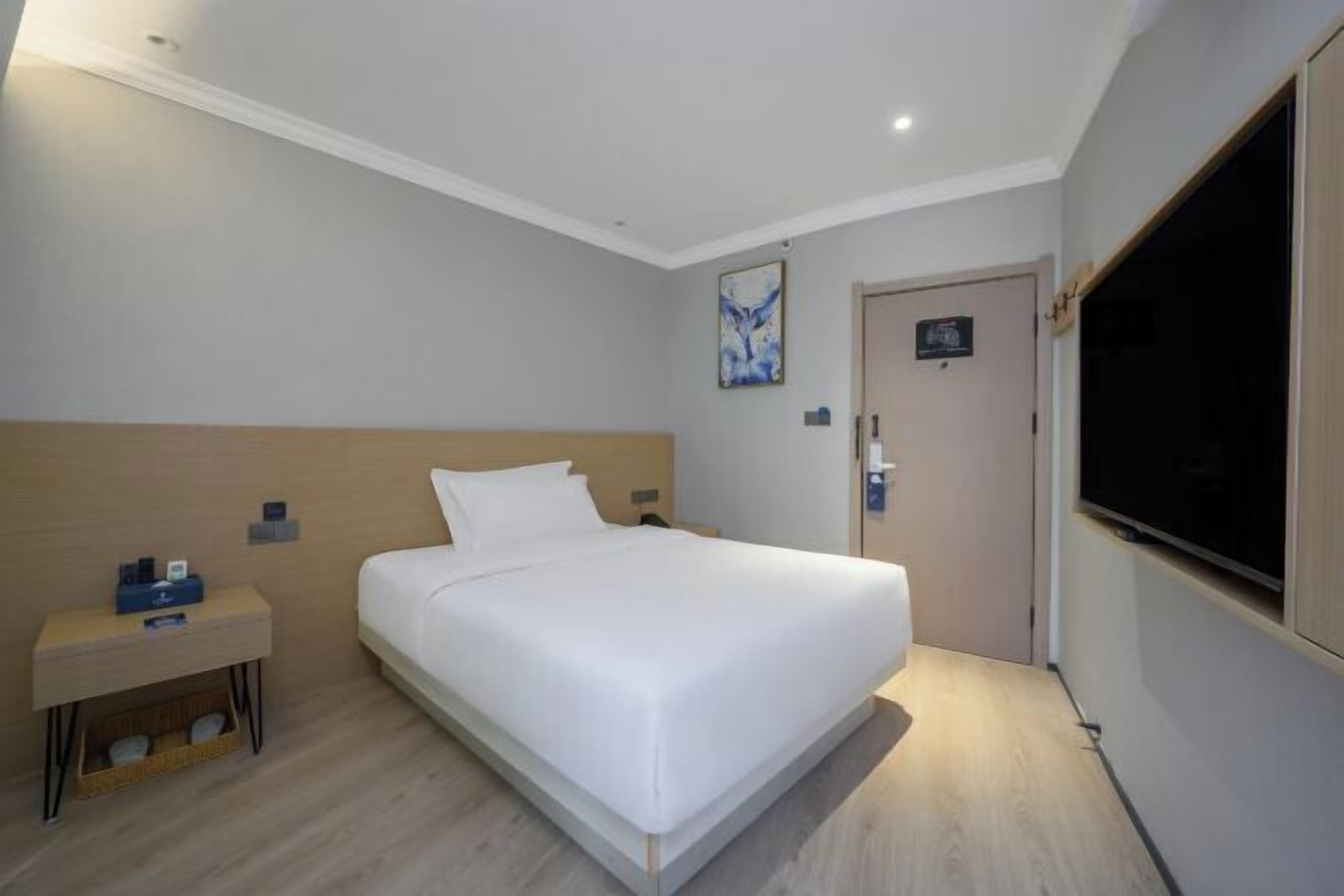 A hotel room with a single bed, a bedside table with a lamp, a TV mounted on the wall, and a door with a sign.