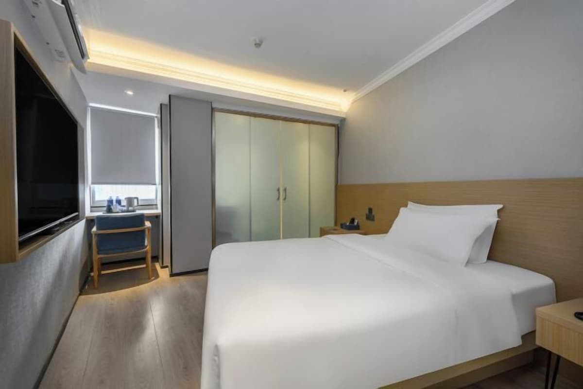 A hotel room with a large bed, a TV mounted on the wall, a desk with a chair, a wardrobe, and a sliding door closet.
