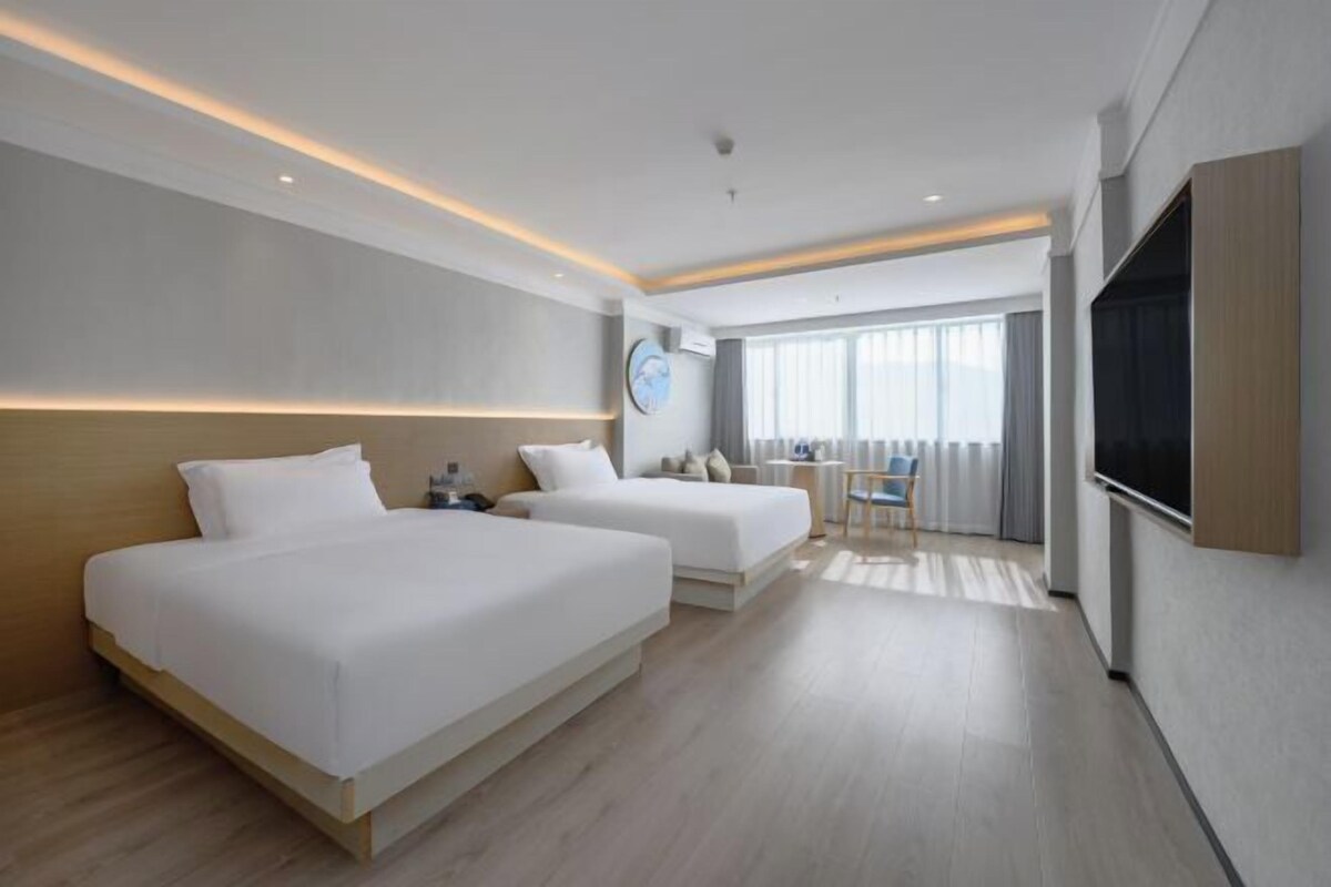 A modern hotel room with two beds, a desk, a chair, a TV, and a large window with curtains.