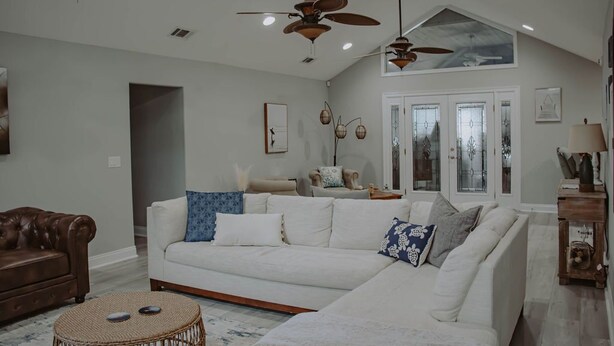 A spacious living room with a large white sectional sofa, a brown leather couch, and a round woven coffee table. There are ceiling fans, recessed lighting, and a large window with a view of trees.