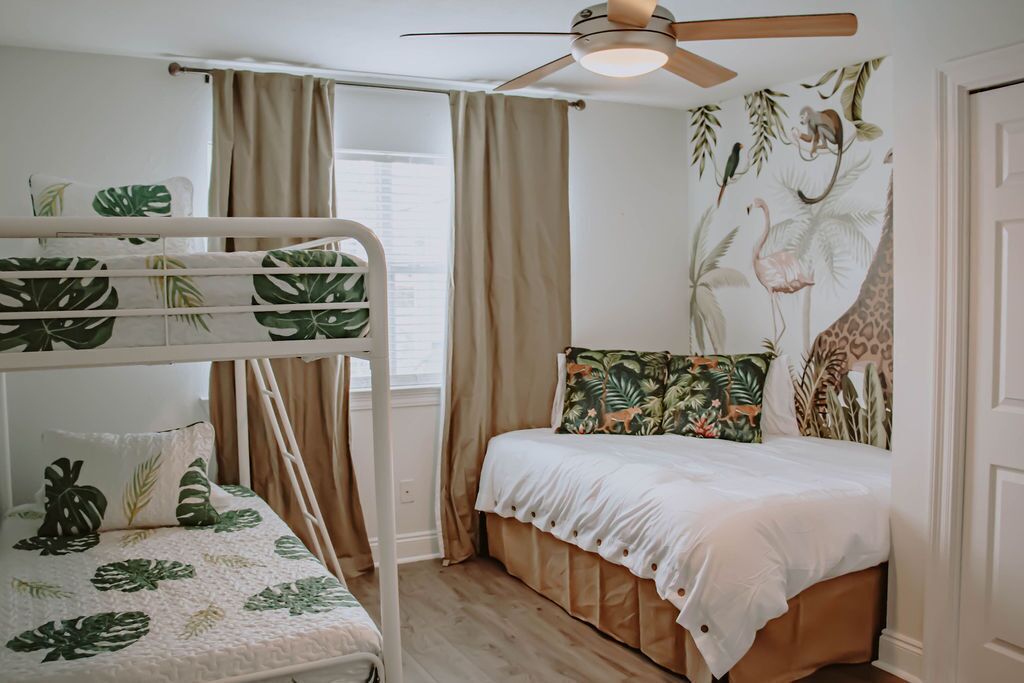 Une chambre avec un lit superposé, un lit simple doté d’une tête de lit à motifs tropicaux, un ventilateur de plafond et une fenêtre avec des rideaux.