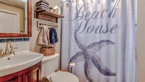 Un bagno con lavandino bianco, mobile contenitore in legno e un water. È presente un appendi asciugamani con asciugamani e uno scaffale sopra il water. Si vede una tenda da doccia con motivo a stella marina e la scritta 'Beach House'.