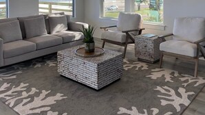 A modern living room with a large ceiling fan, a grey sofa, two armchairs, a coffee table with a plant, and a view of the sea through large windows. - Family-Friendly! (Gulfport)