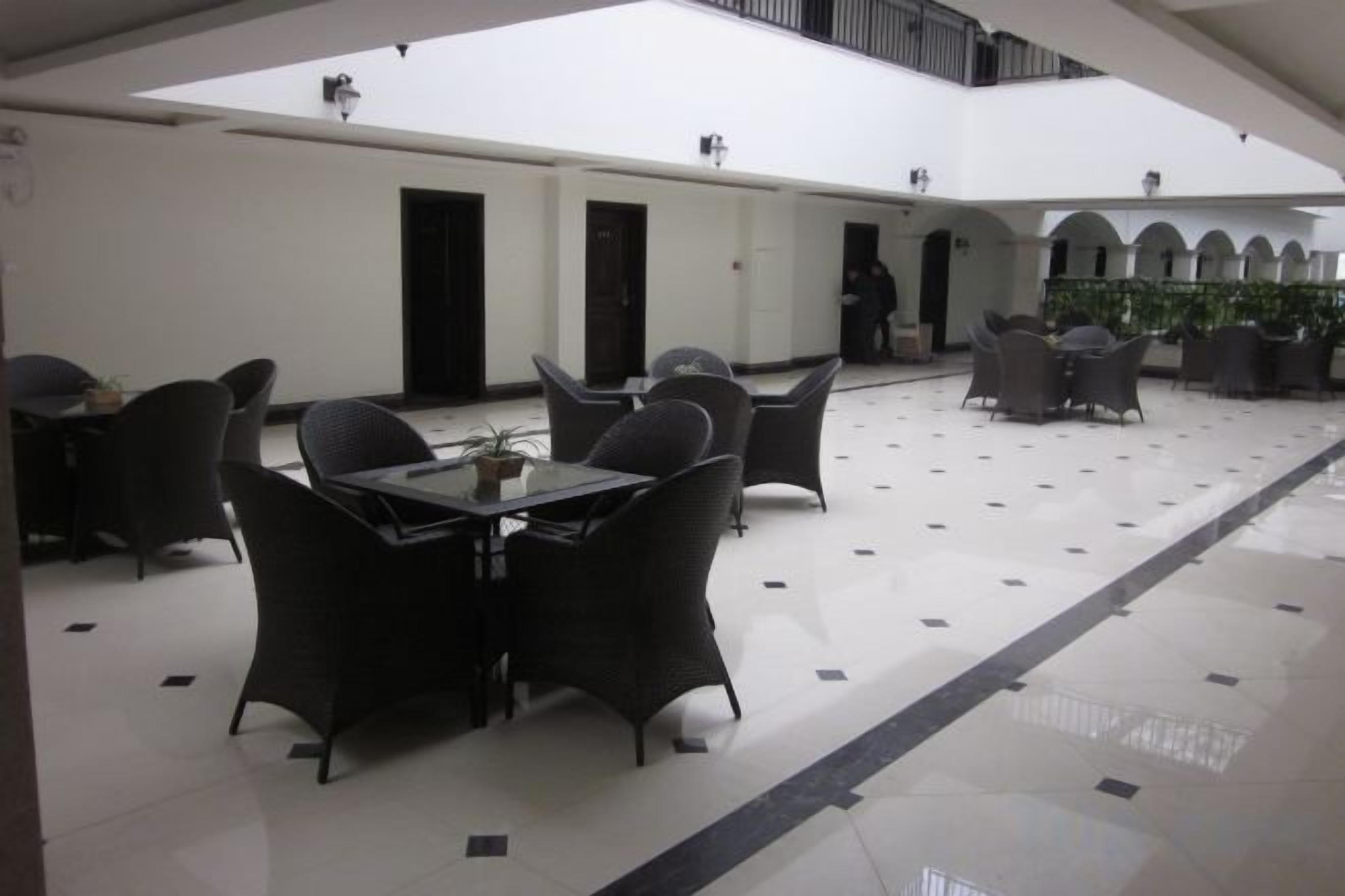 Amplio espacio interior con techo alto, que presenta una zona de estar central con mesas y sillas, rodeada de paredes con puertas y un suelo de baldosas.