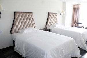 Habitación de hotel con dos camas, cada una con cabecero tapizado, ropa de cama blanca y mesita de noche con lámpara.