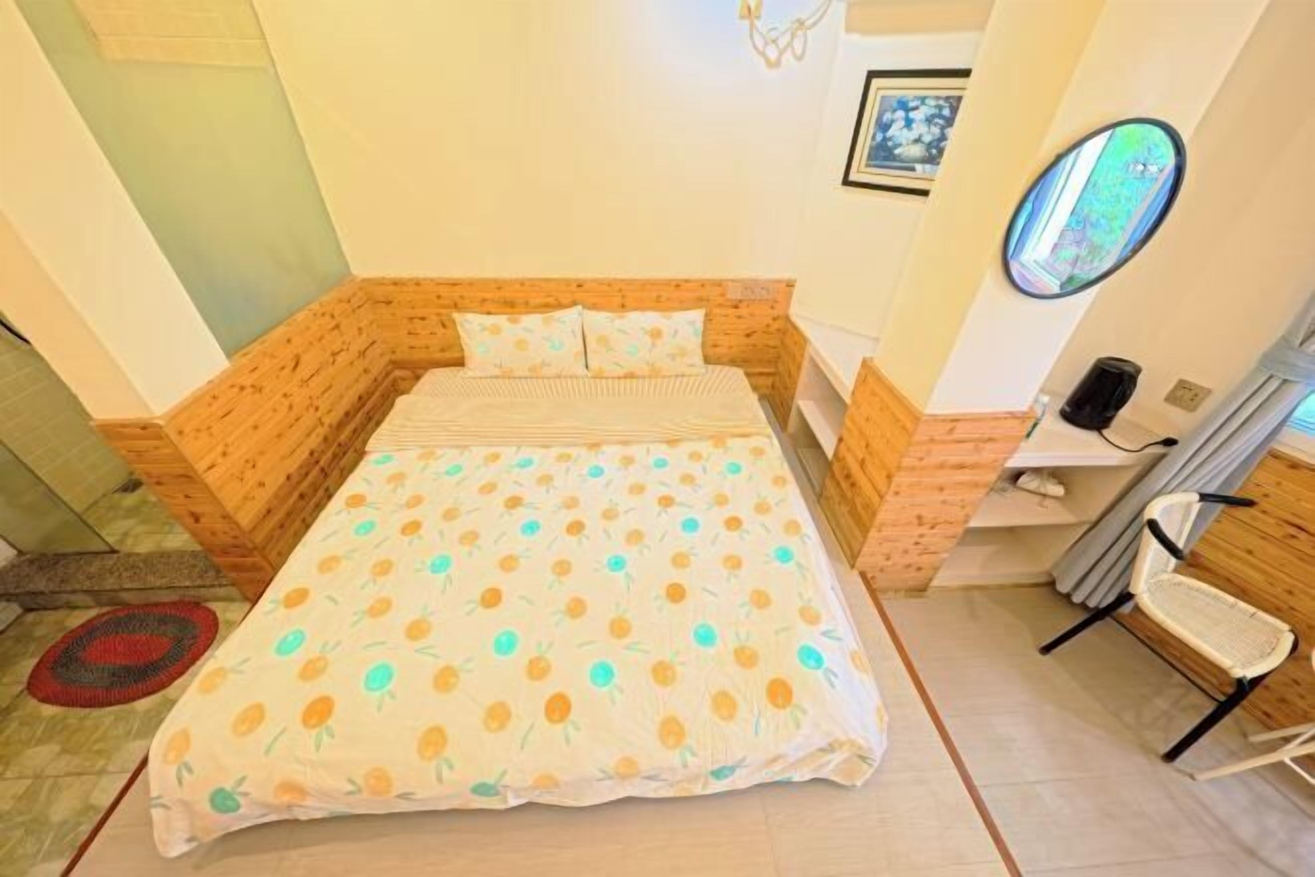 A single bed with a patterned bedspread, a wooden headboard, a small bedside table, a chair, and a mirror on the wall.