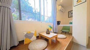 Une pièce avec une grande fenêtre, une petite table en bois, un pouf en osier et une chaise verte.