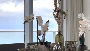 Una mesa de comedor con orquídeas blancas, un florero decorativo y vista al océano a través de amplias ventanas.