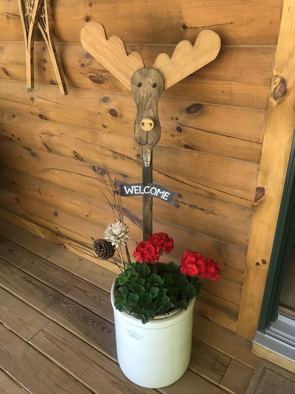 Un panneau en bois orné d’une tête d’élan et d’une inscription « BIENVENUE », une plante en pot avec des fleurs rouges et une pomme de pin.