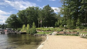 A lakeside area with a sandy beach, a dock with boats, and a row of chairs. - Luxury Log home steps from private beach in Meredith on Lake Winnipesaukee. (Meredith)