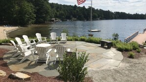 A lakeside patio with Adirondack chairs, a flagpole with an American flag, and a dock with boats. - Luxury Log home steps from private beach in Meredith on Lake Winnipesaukee. (Meredith)