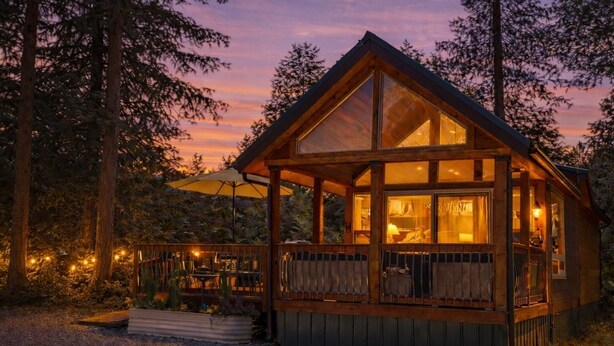 A wooden cabin with a large glass window, a deck with outdoor seating, and a view of the surrounding forest. - Magical Forest Cabin- Secluded Retreat Near Beach (Lakebay)