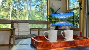 A room with a wooden bench, two white mugs on a tray, a TV displaying a mountain landscape, and a large window with a view of trees. - Magical Forest Cabin- Secluded Retreat Near Beach (Lakebay)