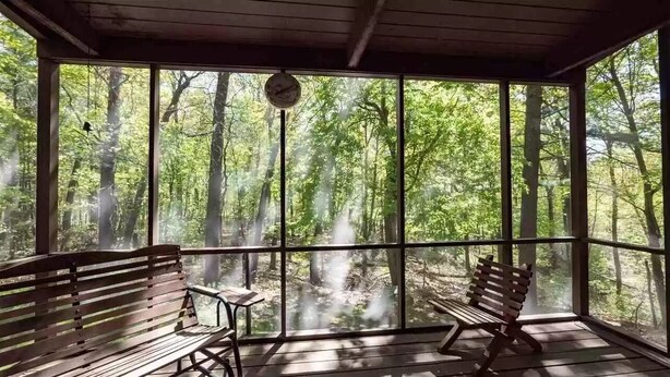 A covered porch with two wooden benches and a small table, overlooking a forested area.